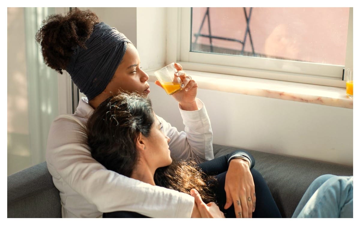 Mujer tomando un jugo mientras abraza a otra persona en un ambiente cálido y tranquilo junto a una ventana, reflejando calma y conexión emocional.