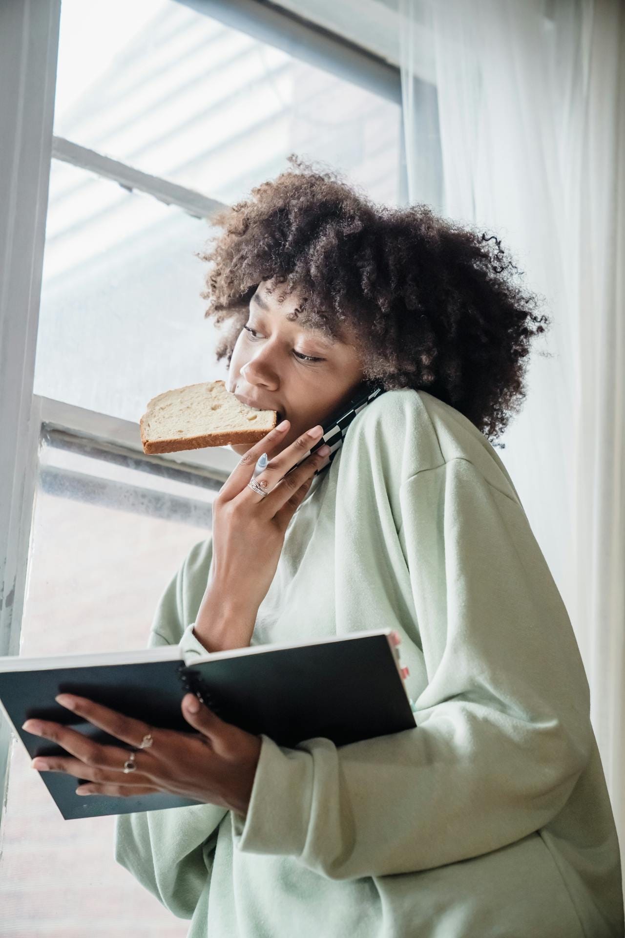 Mujer comiendo, sostendiendo el teléfono con una mano, mirando por la ventana y sosteniendo una libreta con la otra mano.